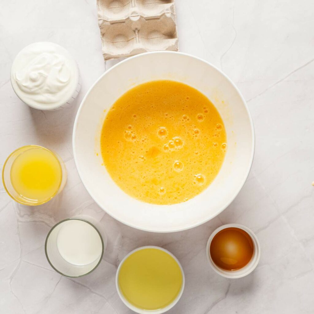 A white bowl with beaten eggs surrounded by a glass of milk, a cup of oil, yogurt, juice, and an egg on a marble surface.