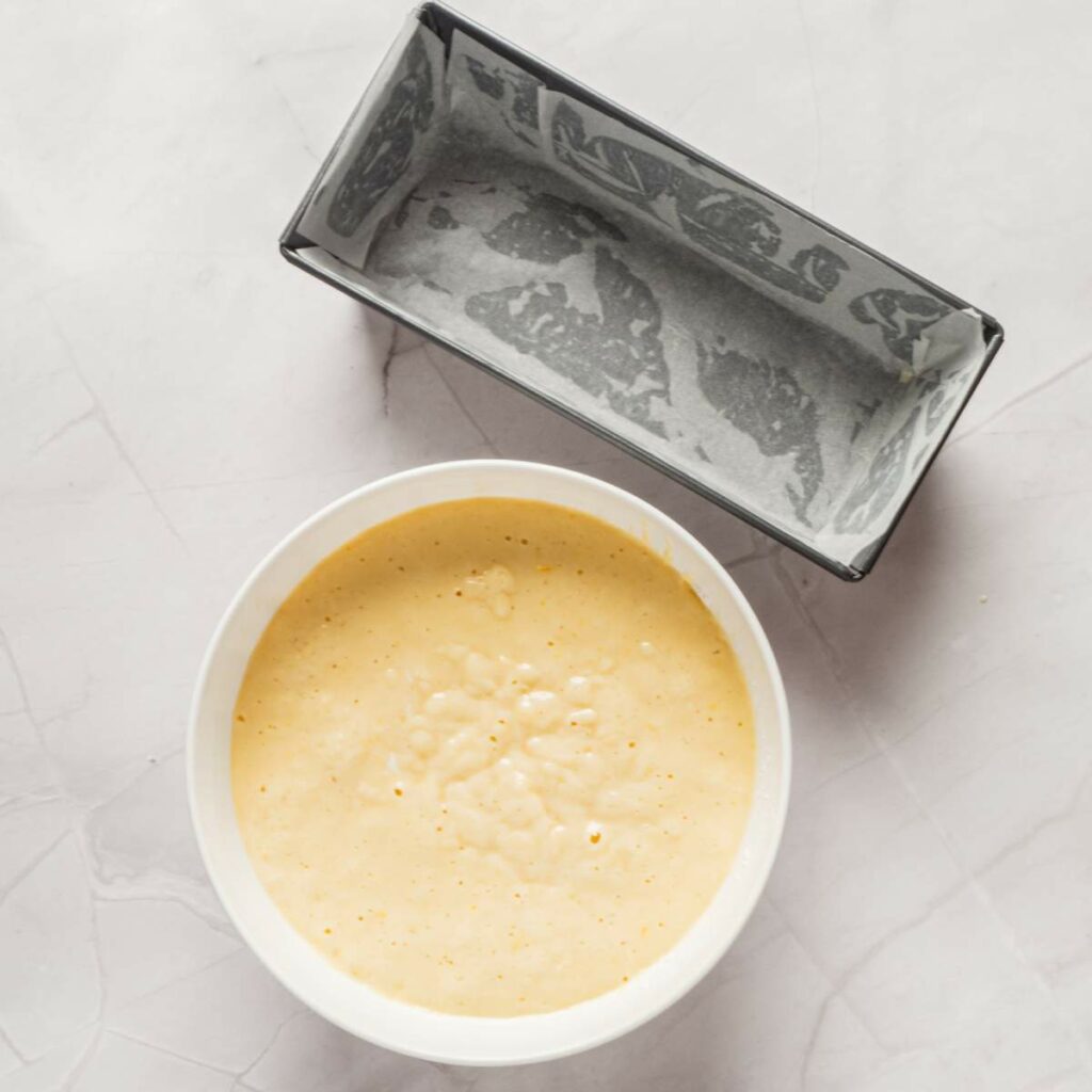 A bowl of light yellow cake batter is beside a lined, rectangular baking pan on a gray surface.