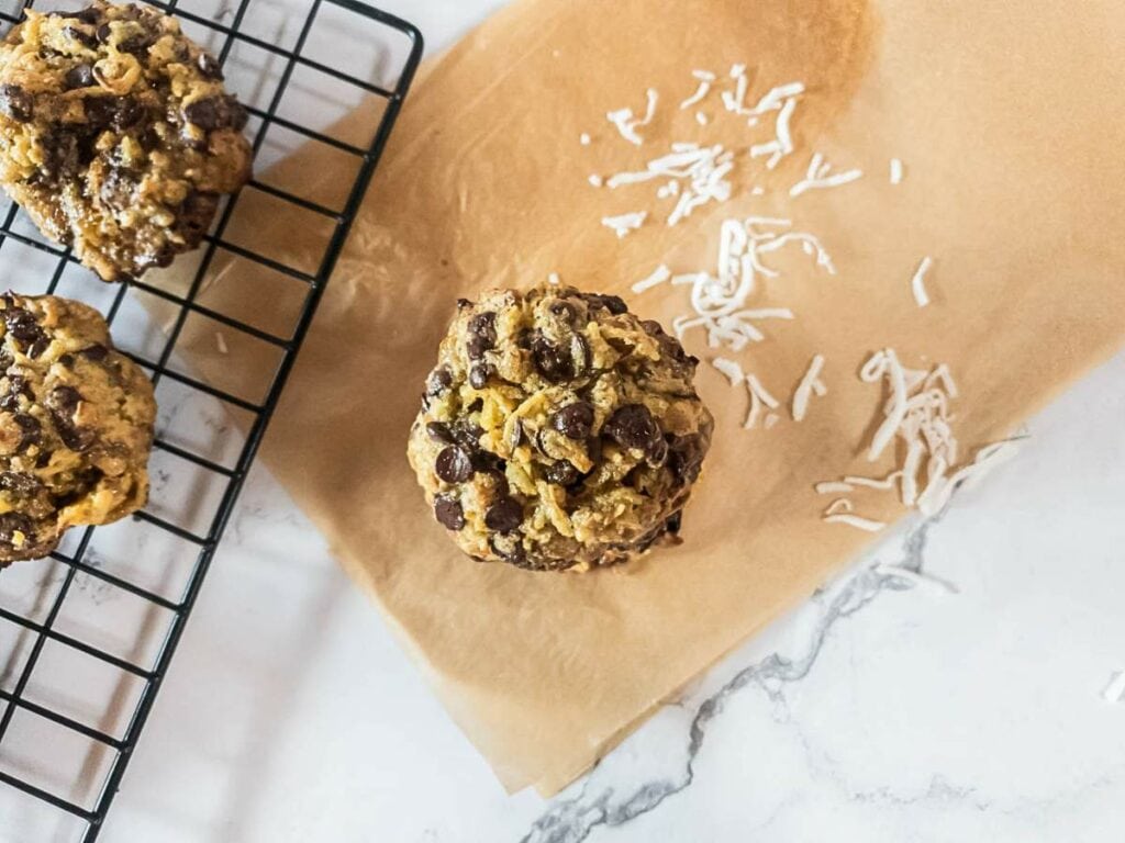 Magic cookie on parchment paper with coconut shreds beside it, cooling rack with other cookies on the left, set on a marble surface.