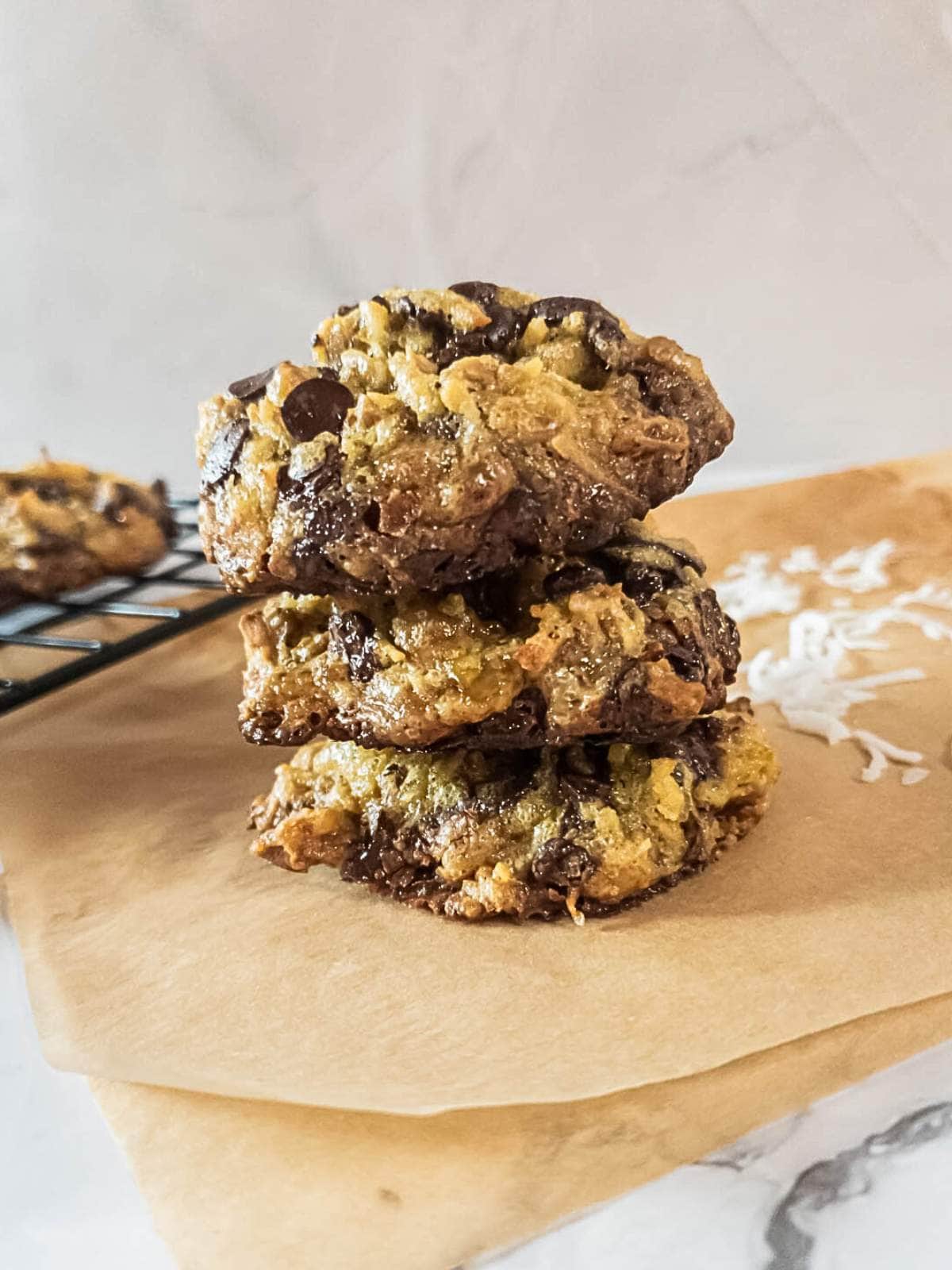 Three magic cookies stacked on a piece of parchment paper with another cookie on a cooling rack in the background.