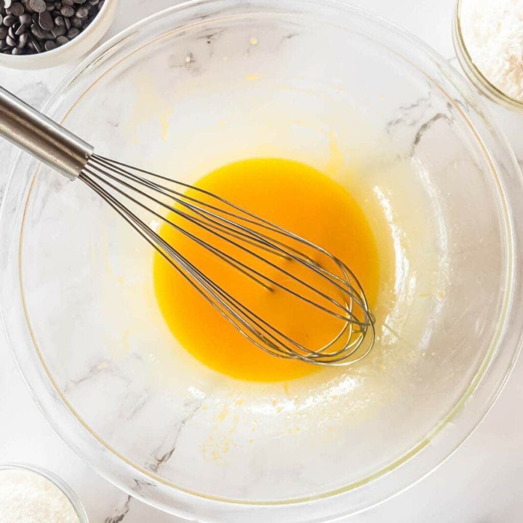 A whisk in a glass bowl with yellow liquid batter. Chocolate chips and flour are in bowls nearby on a marble surface.