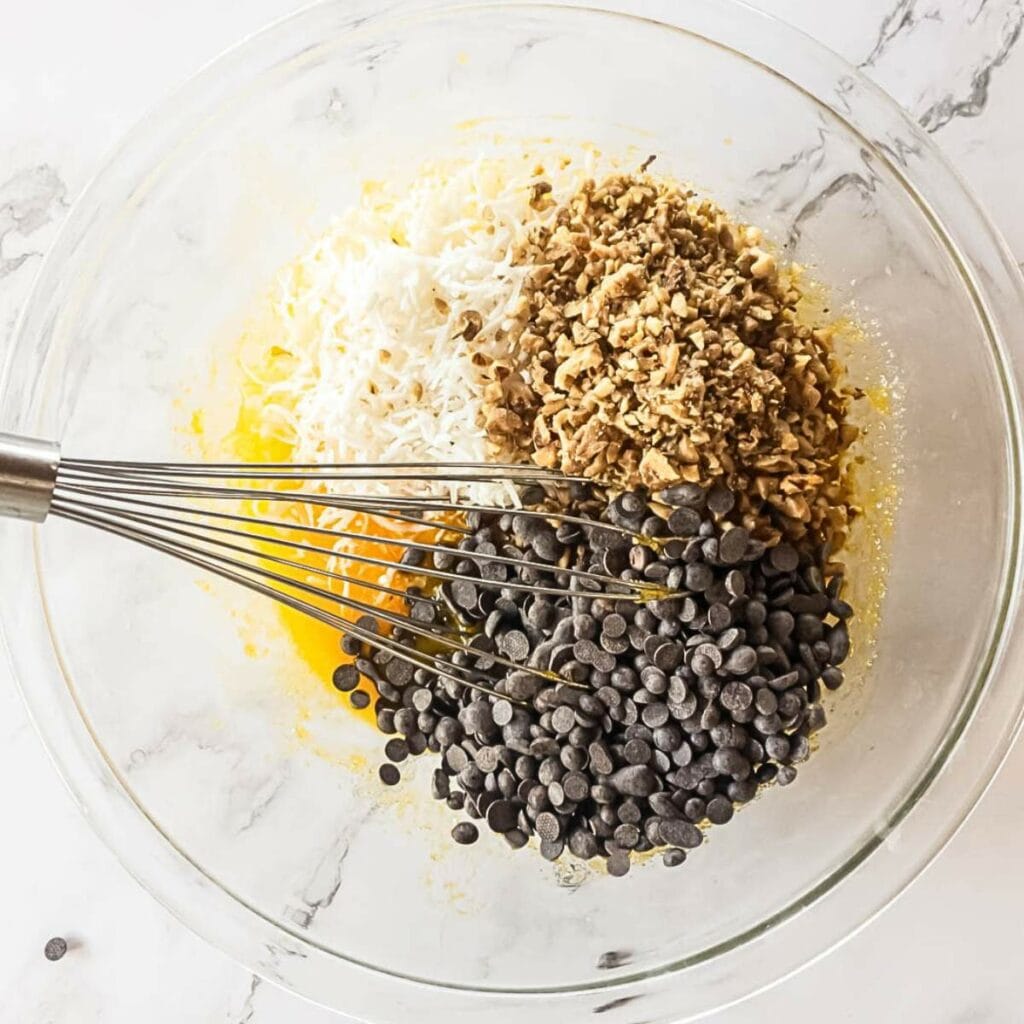 Mixing bowl with chocolate chips, chopped nuts, shredded coconut, and egg mixture, with a whisk on marble surface.