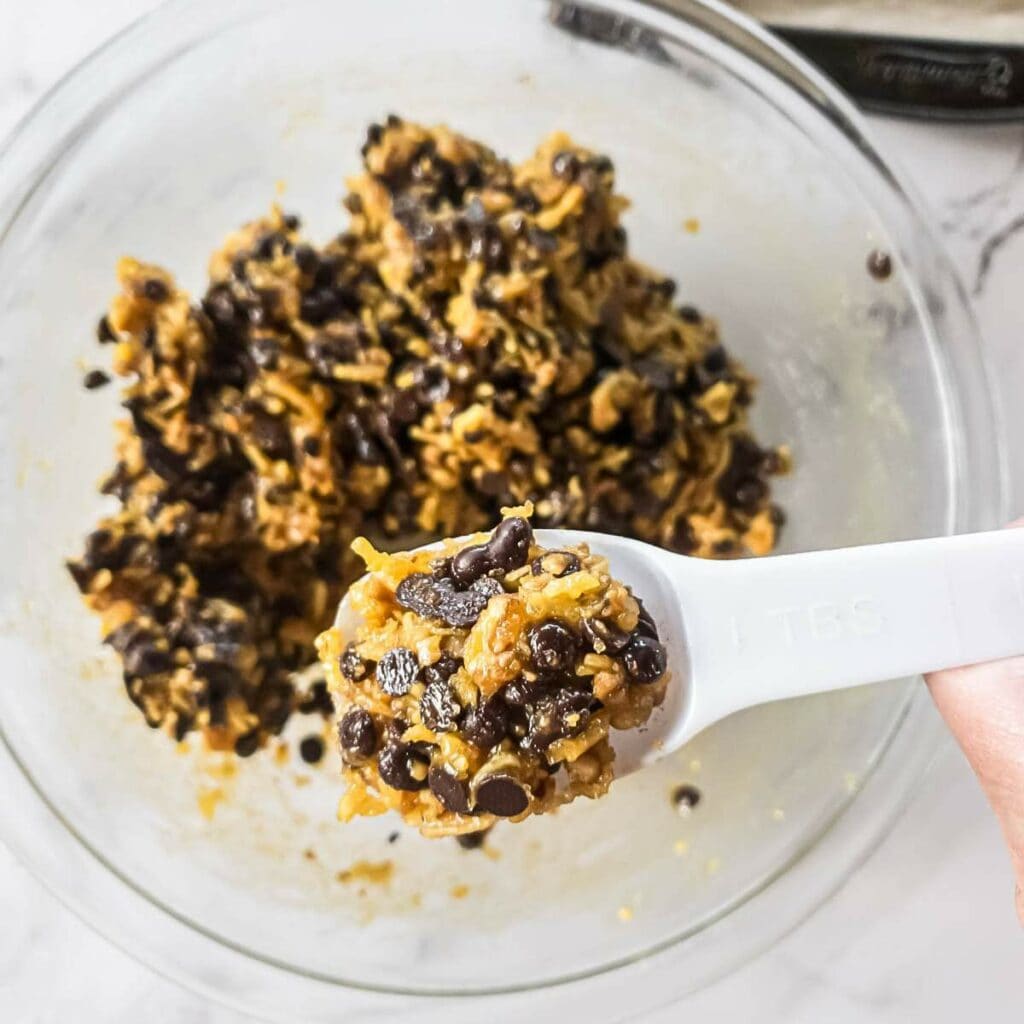 Close-up of a tablespoon scooping cookie dough with chocolate chips from a glass bowl on a marble countertop.