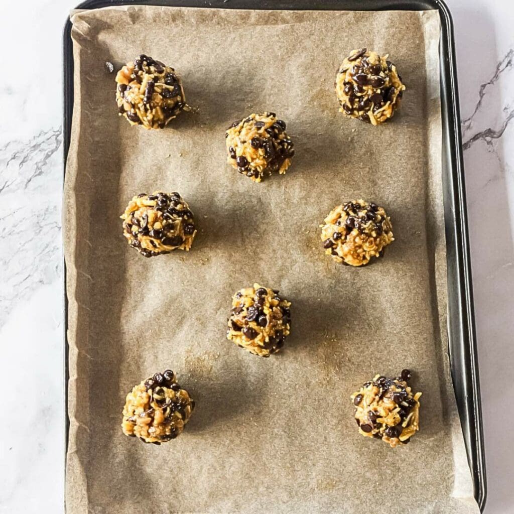 Eight unbaked cookie dough balls with chocolate chips and nuts on a parchment-lined baking tray.