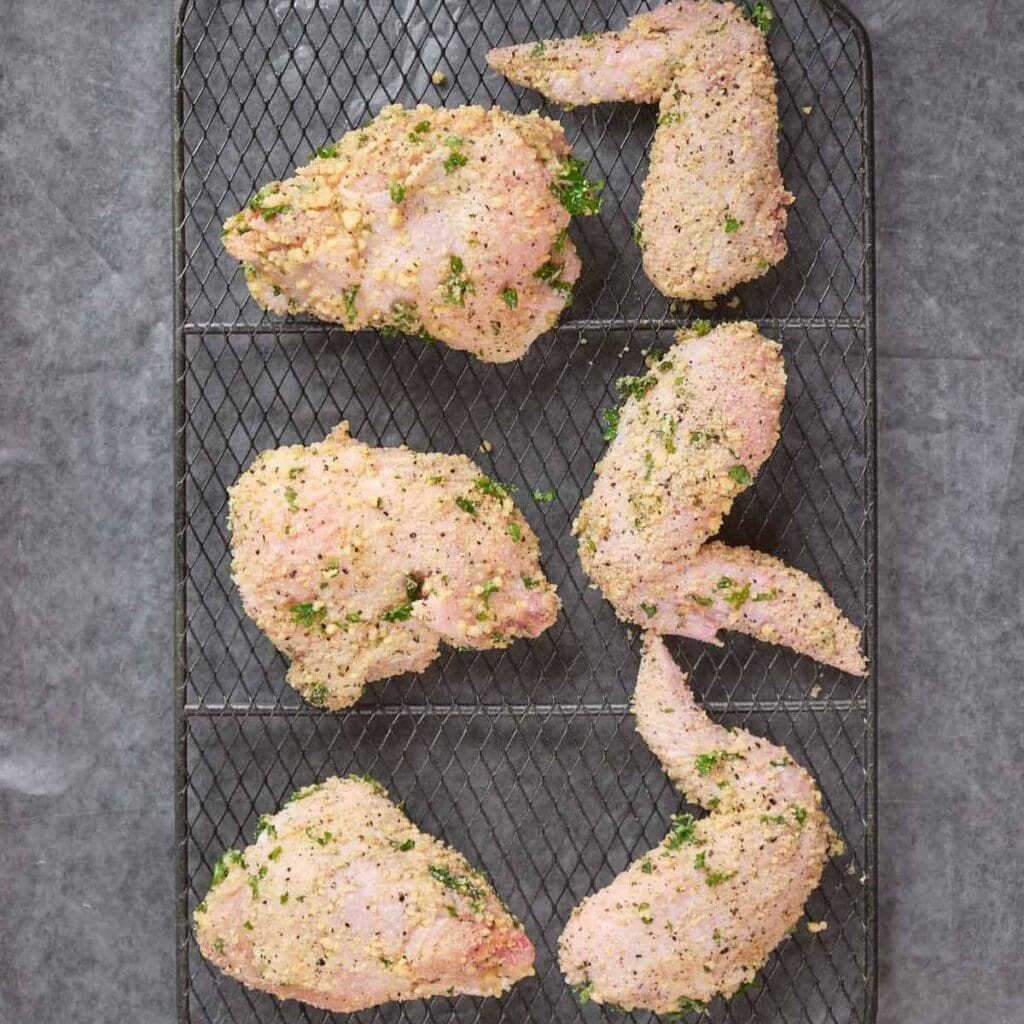 Raw breaded chicken pieces with herbs are arranged on a wire rack over a grey surface, ready for cooking.