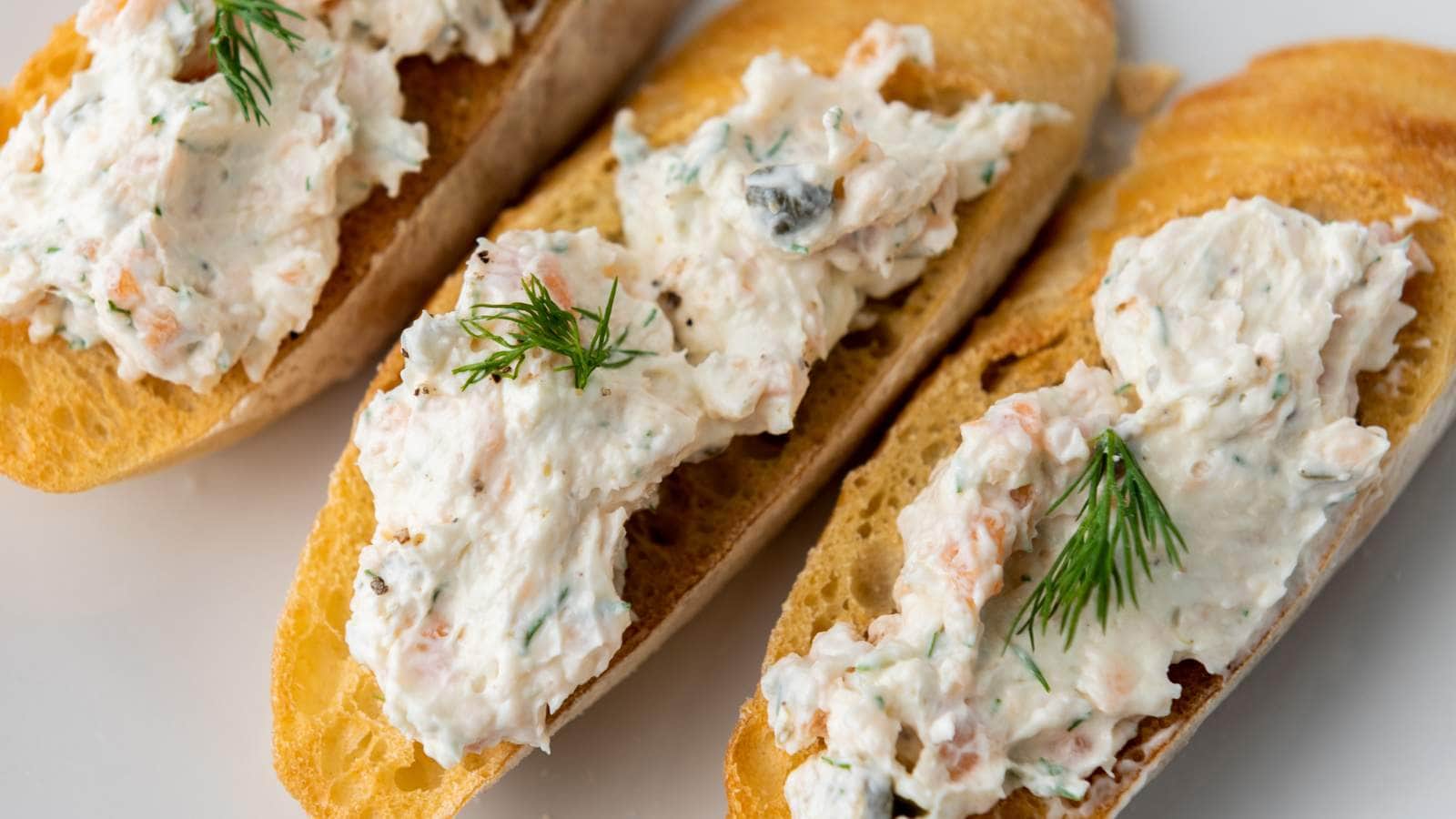 Three slices of toasted baguette topped with a smoked salmon dip garnished with fresh dill on a white surface.