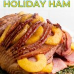 A sliced holiday ham garnished with pineapple rings and herbs, displayed on a wooden board with the text "Juicy Pineapple Holiday Ham" above it.