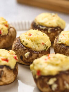 Close-up of stuffed mushrooms topped with a creamy, herb-infused cheese mixture and small red pepper pieces, arranged on a white plate.