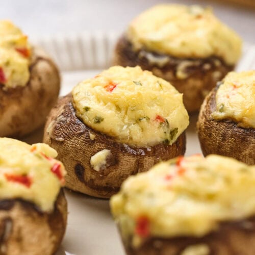 Close-up of stuffed mushrooms topped with a creamy, herb-infused cheese mixture and small red pepper pieces, arranged on a white plate.