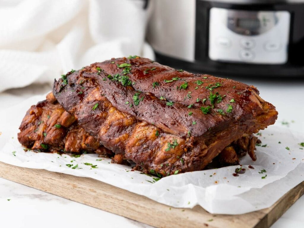 A rack of cooked baby back ribs garnished with chopped herbs sits on parchment paper atop a wooden board.
