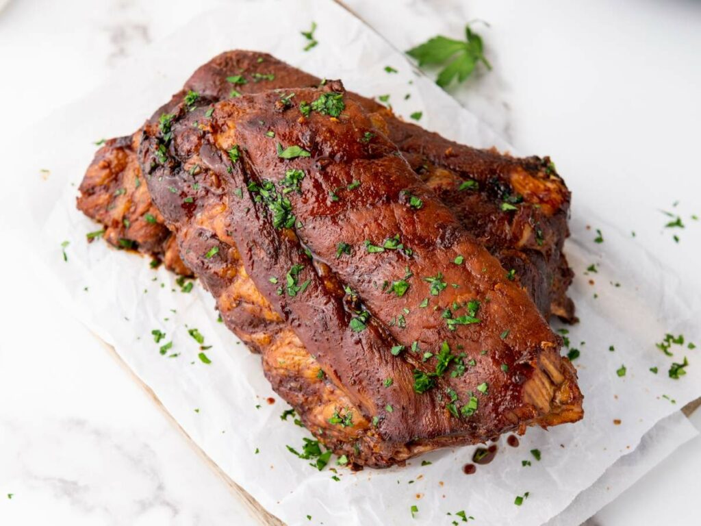 Baby back ribs stacked on parchment paper, garnished with chopped parsley, on a white marble surface.