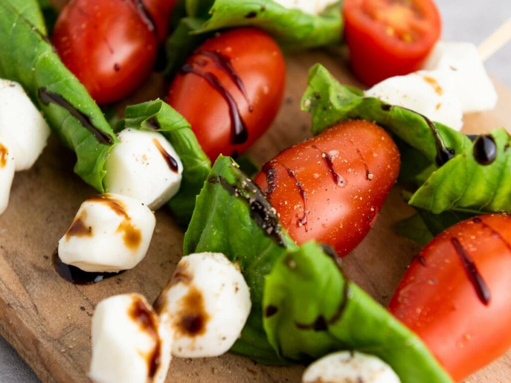 Close-up of skewered cherry tomatoes, mozzarella balls, and basil leaves on a wooden board, drizzled with balsamic glaze.