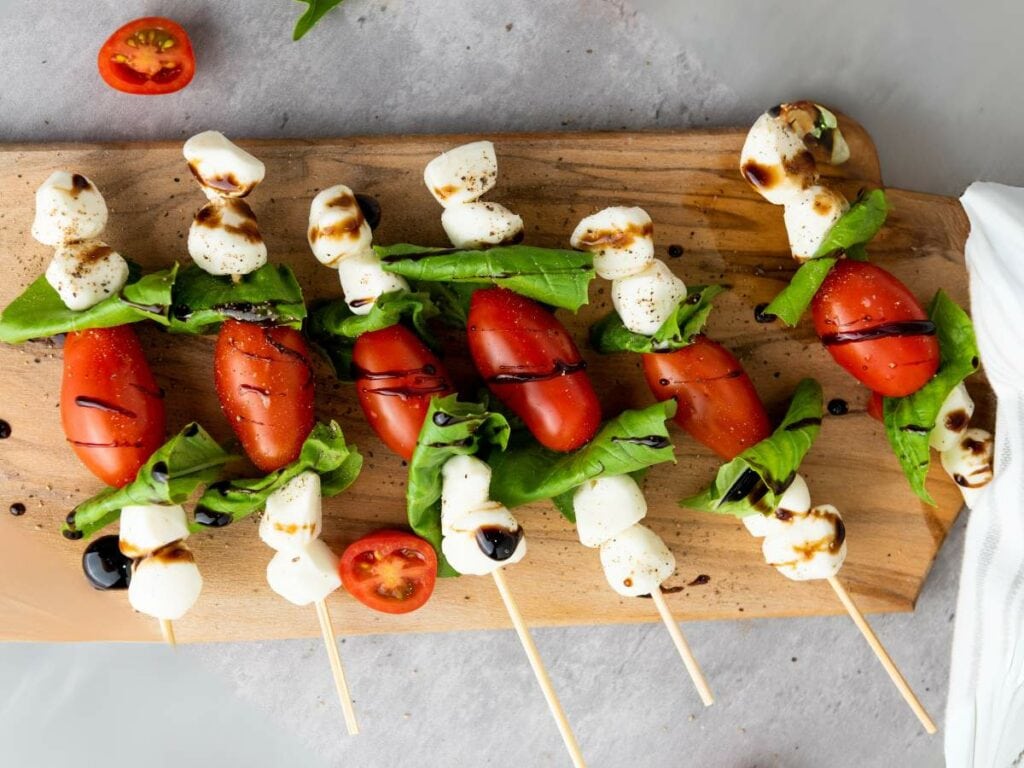 Skewers with cherry tomatoes, mozzarella balls, and basil leaves drizzled with balsamic glaze on a wooden board.