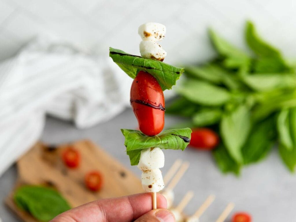Hand holding a skewer with cherry tomato, mozzarella balls, and basil leaves.
