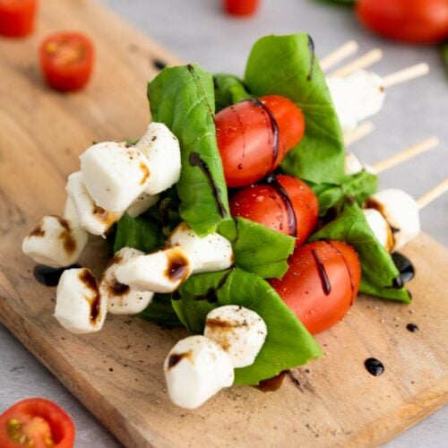 Skewers with cherry tomatoes, mozzarella balls, and basil leaves on a wooden board, drizzled with balsamic glaze.
