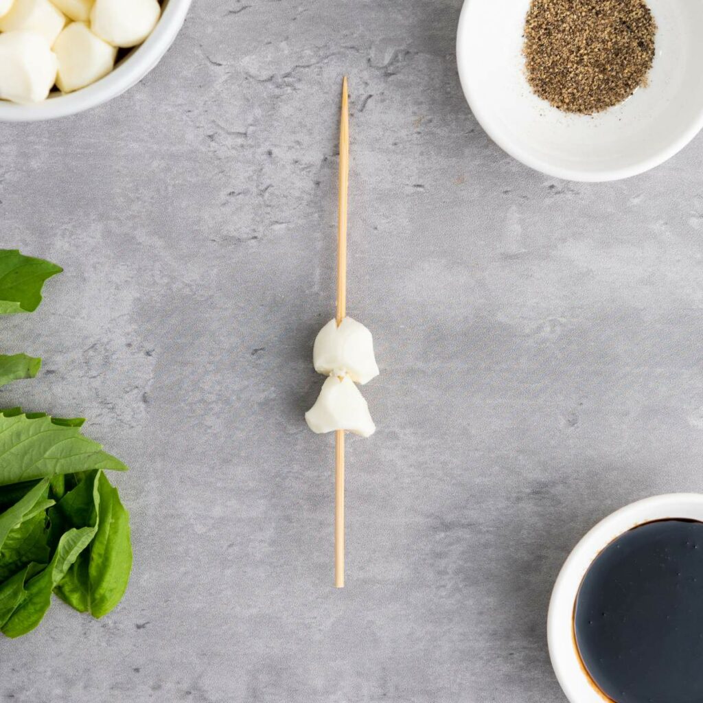 A skewer with mozzarella balls on a gray surface, surrounded by bowls of mozzarella, black pepper, balsamic vinegar, and fresh basil leaves.