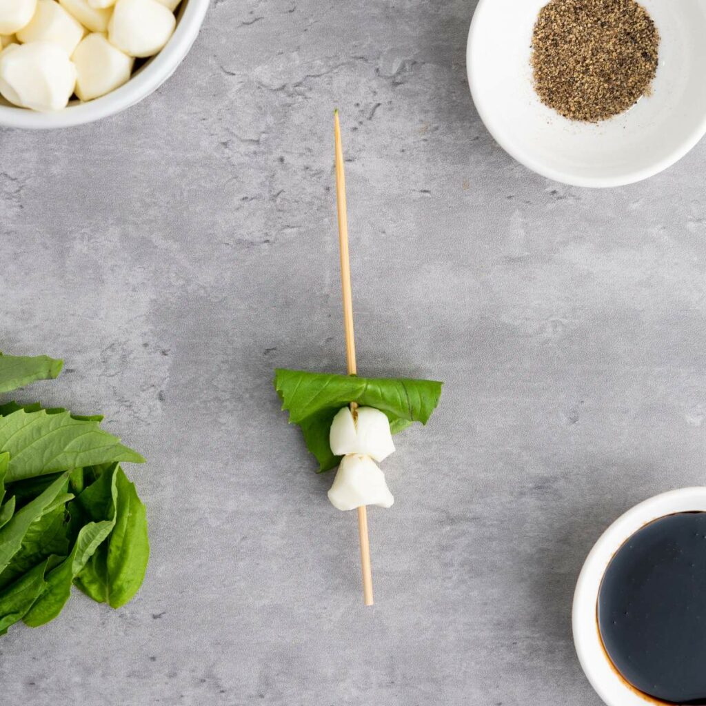 A skewer with two mozzarella balls and a basil leaf on a gray surface.
