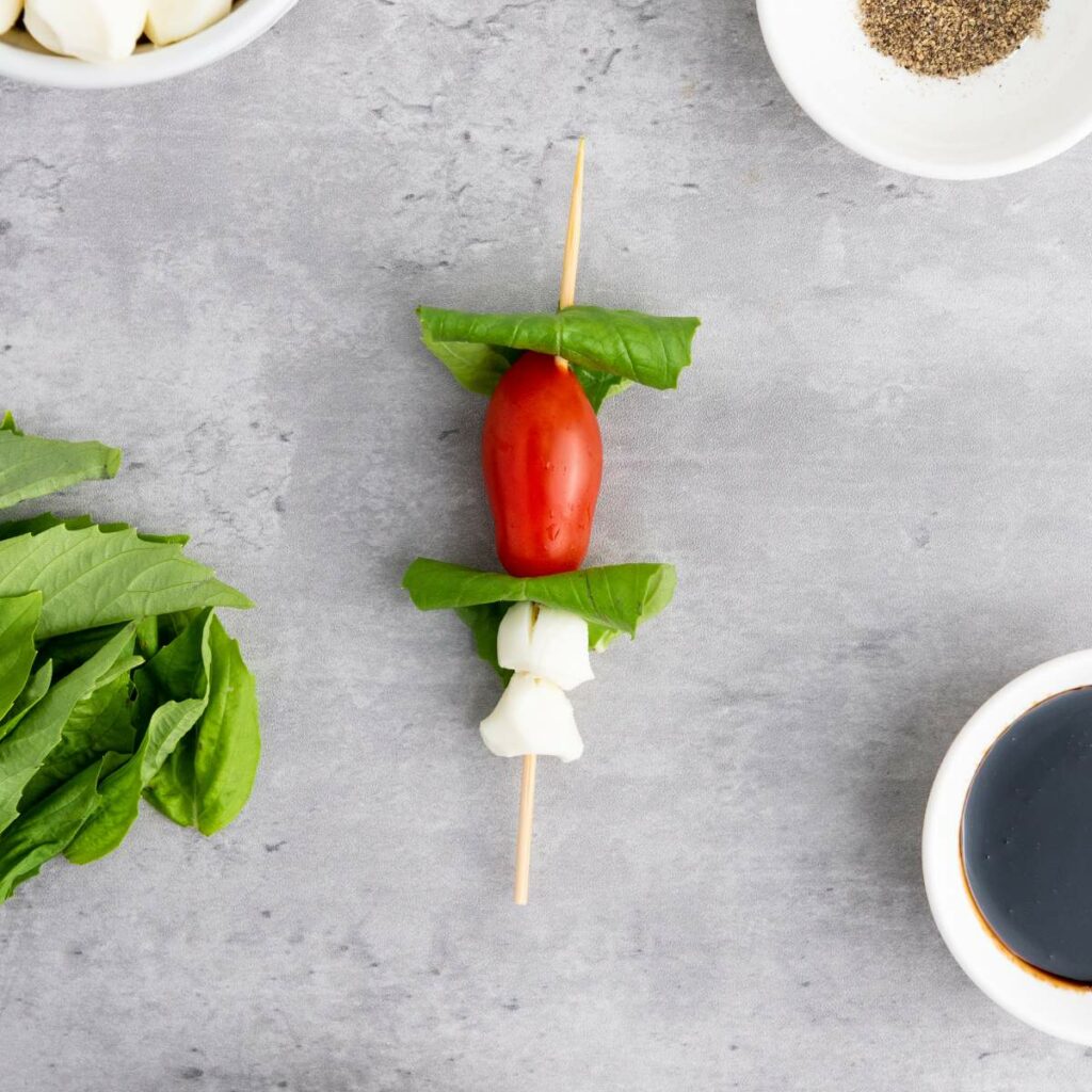 Skewer with a cherry tomato, basil leaves, and mozzarella, surrounded by bowls of ingredients on a gray surface.