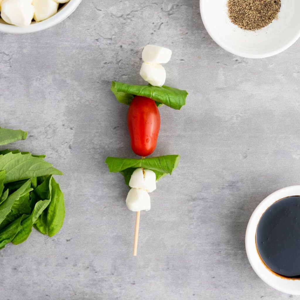 Skewer with cherry tomato, mozzarella balls, and basil leaves on a gray surface, surrounded by bowls of pepper, sauce, cheese, and basil.