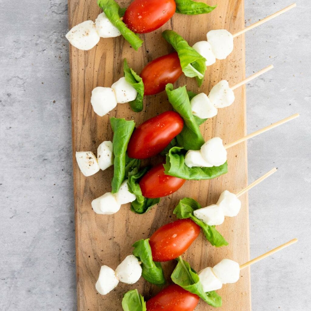 Skewers with cherry tomatoes, mozzarella balls, and basil leaves on a wooden board.
