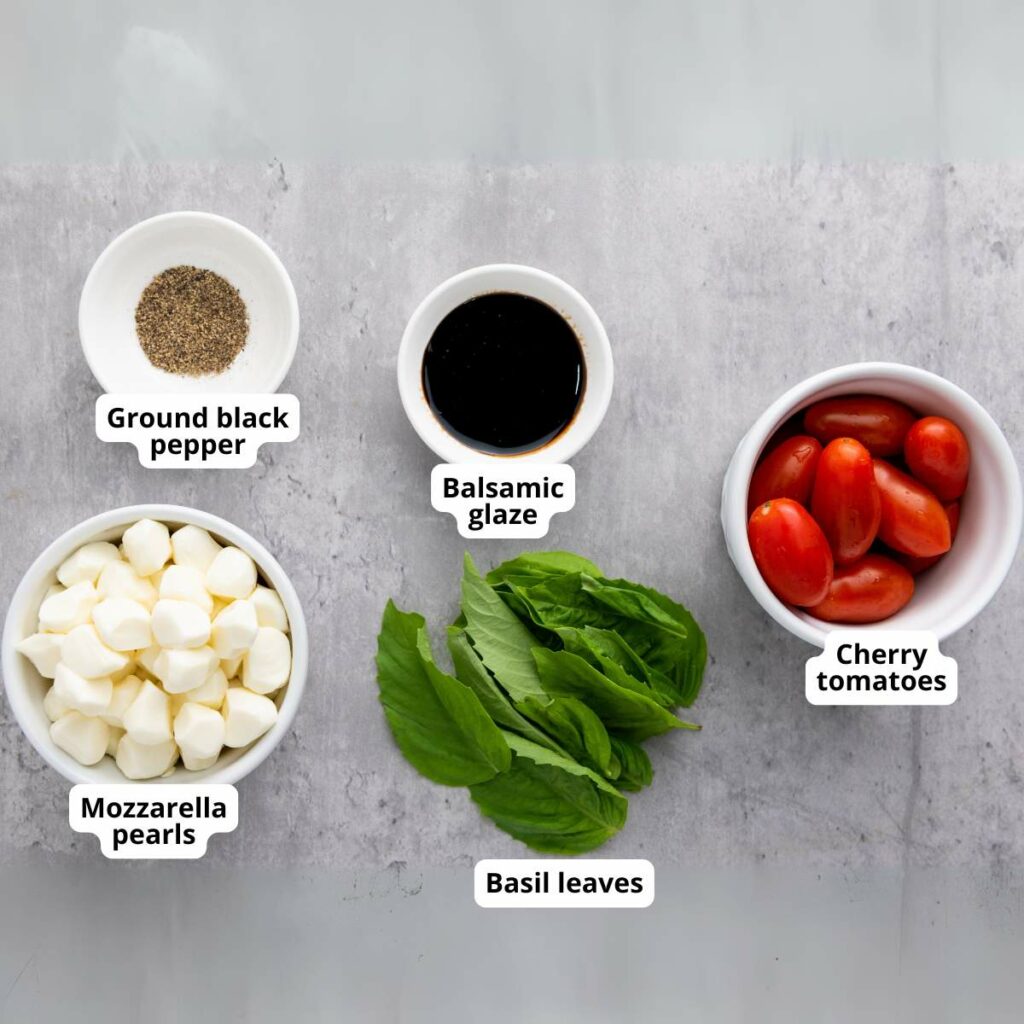Bowls of ground black pepper, balsamic glaze, cherry tomatoes, mozzarella pearls, and fresh basil leaves arranged on a gray surface.