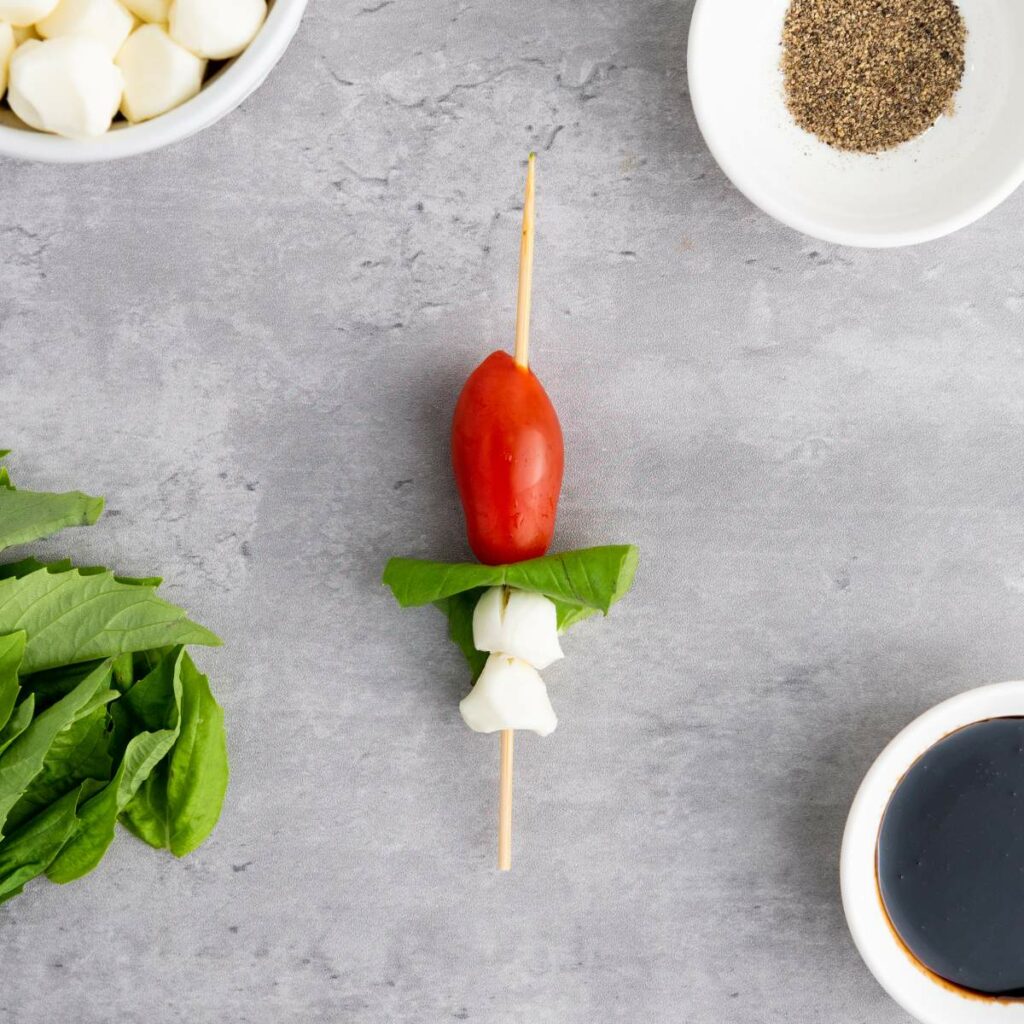 A skewer with a cherry tomato, mozzarella, and basil leaf on a gray surface.