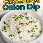 A bowl of creamy caramelized onion dip topped with chopped chives and black pepper, with part of a bowl of round crackers visible in the background.