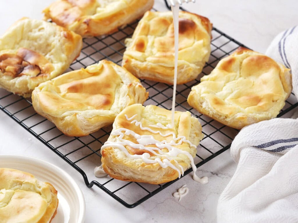 Cheese danish cooling on a wire rack, with icing being drizzled onto one of them.