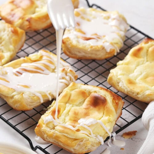 A fork drizzles white icing over cheese danish on a wire cooling rack.