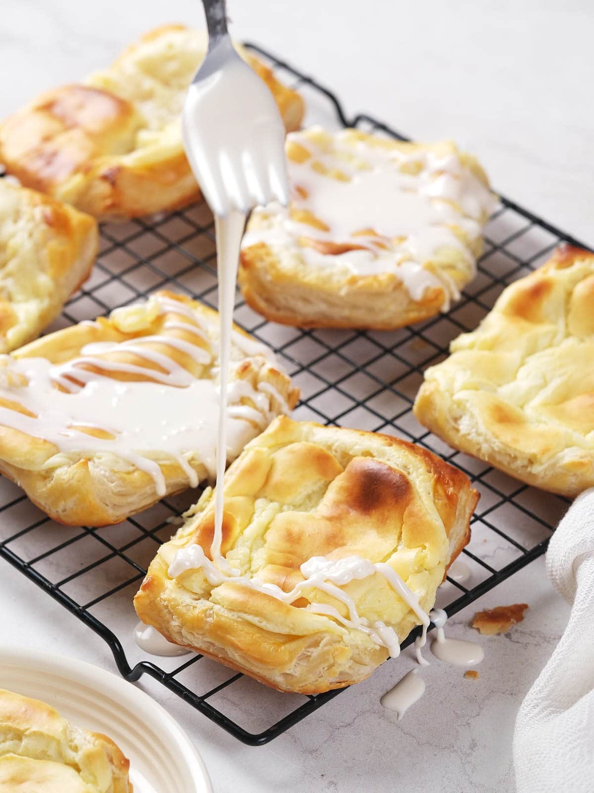 A fork drizzles white icing over cheese danish on a wire cooling rack.