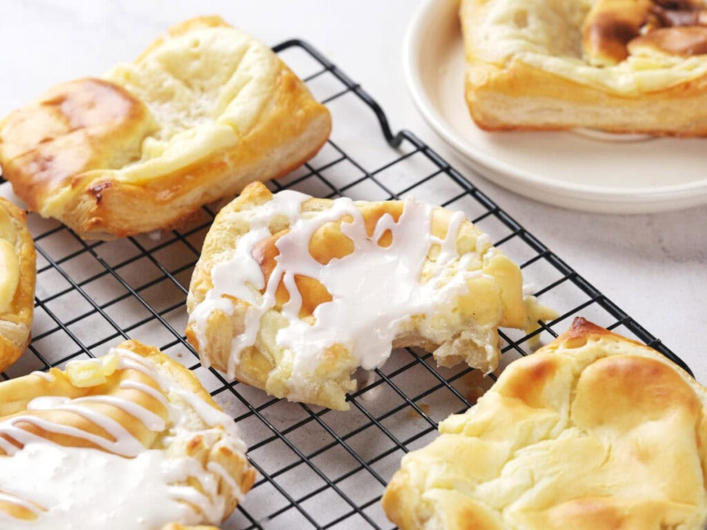 Cheese danish with icing on a cooling rack, one with a bite taken out, with more pastries on a nearby plate.