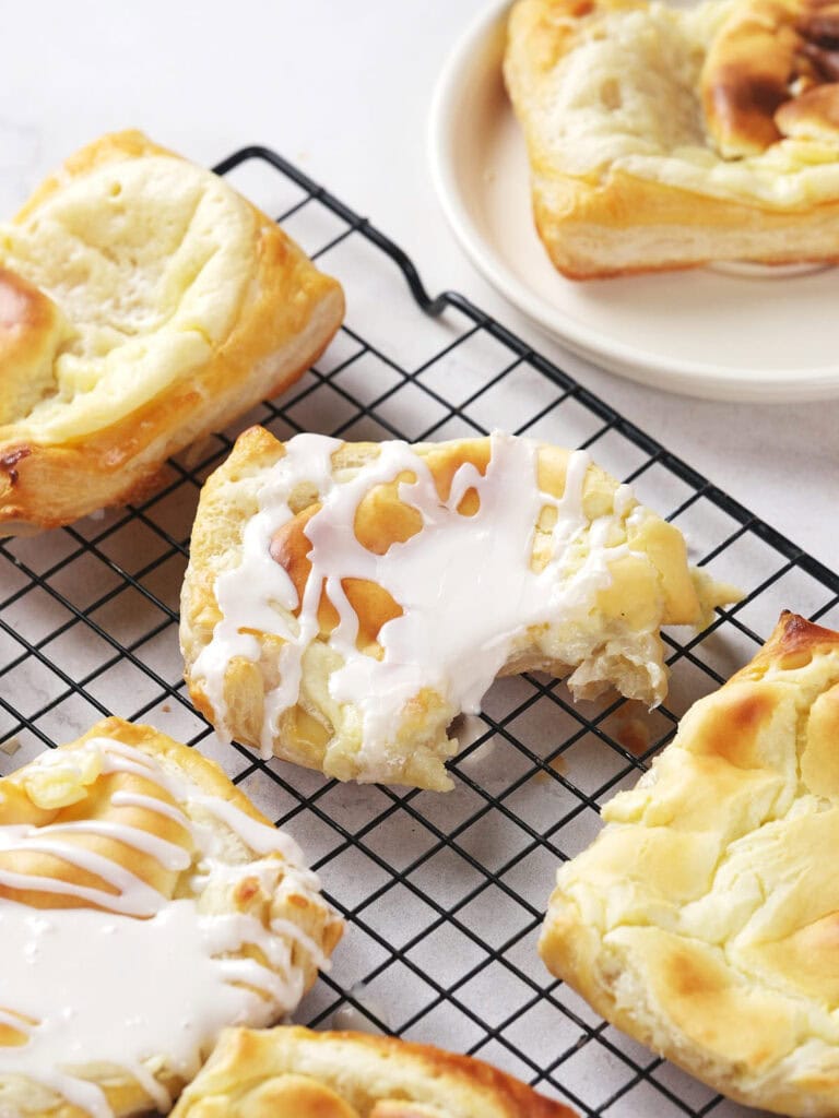 Several cheese danish with white icing are cooling on a black wire rack, with one partially eaten.
