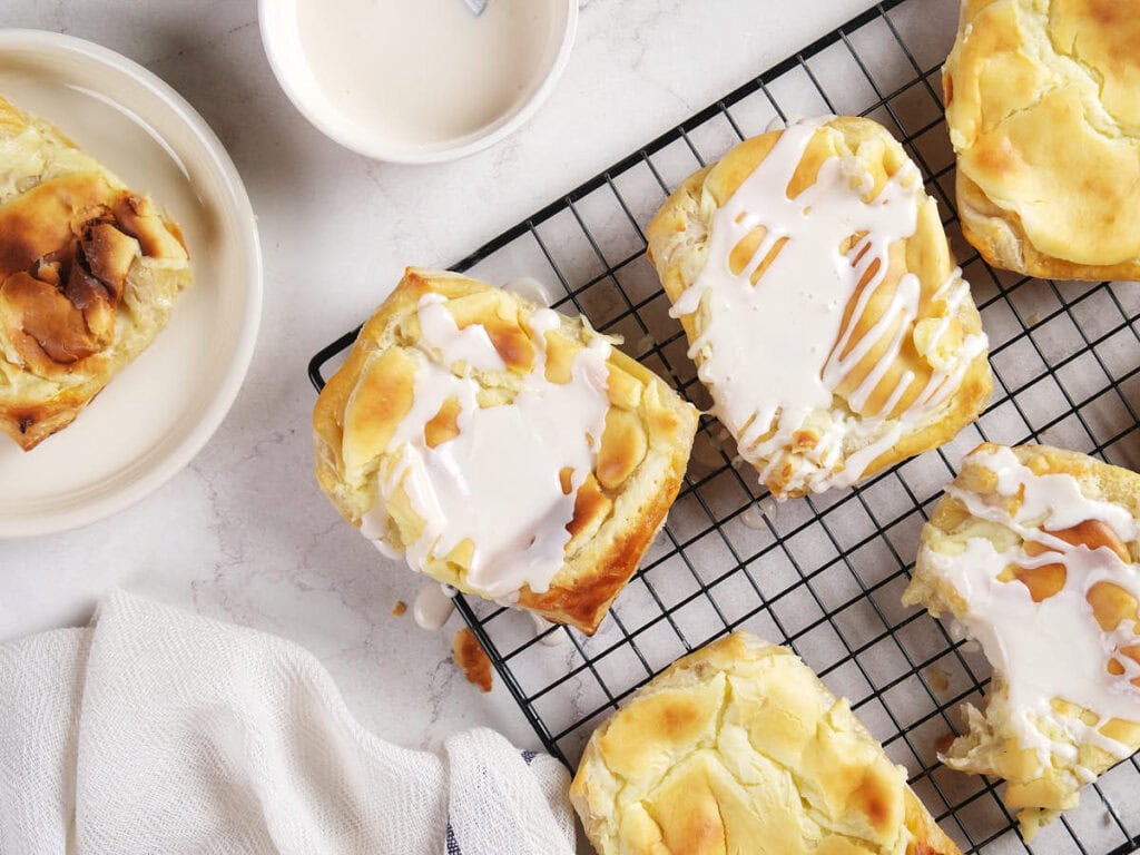 Cheese danish on a black cooling rack, with a partially eaten piece and a bowl of icing nearby on a white surface.