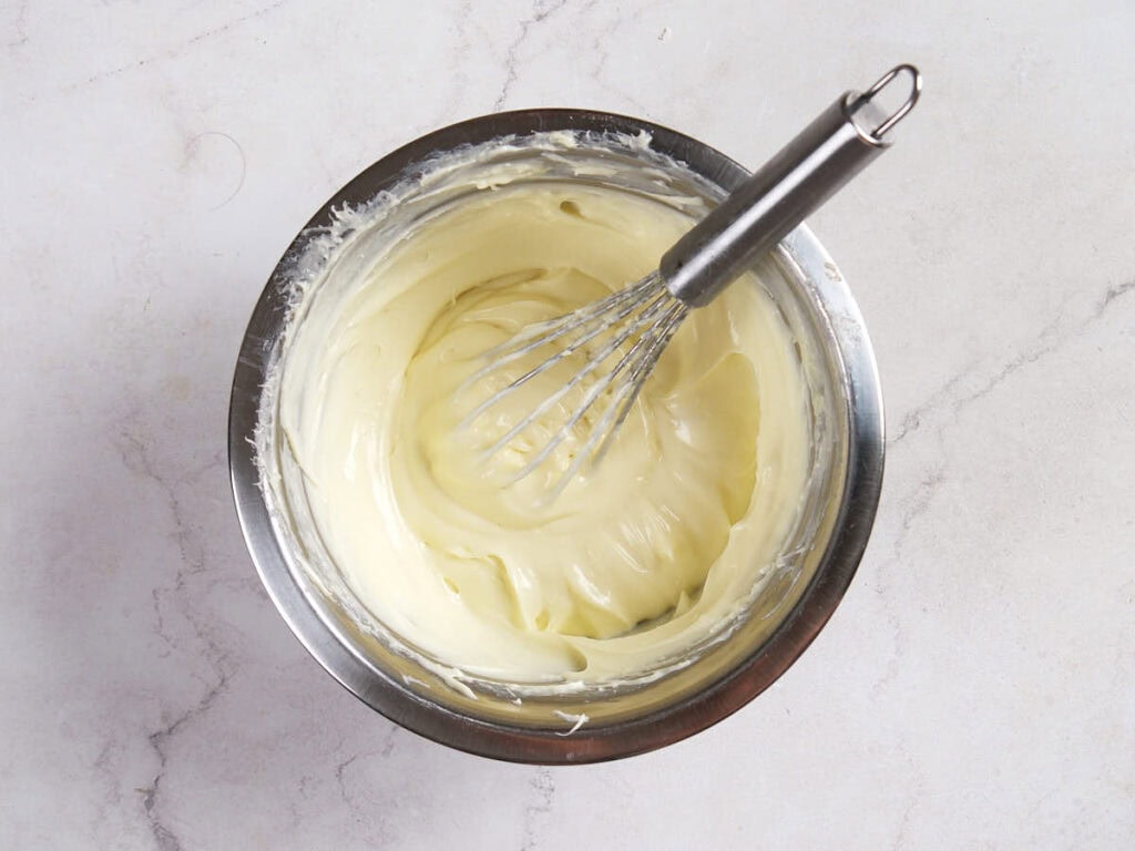 A metal bowl containing creamy, pale batter with a metal whisk resting inside, placed on a light marble surface.