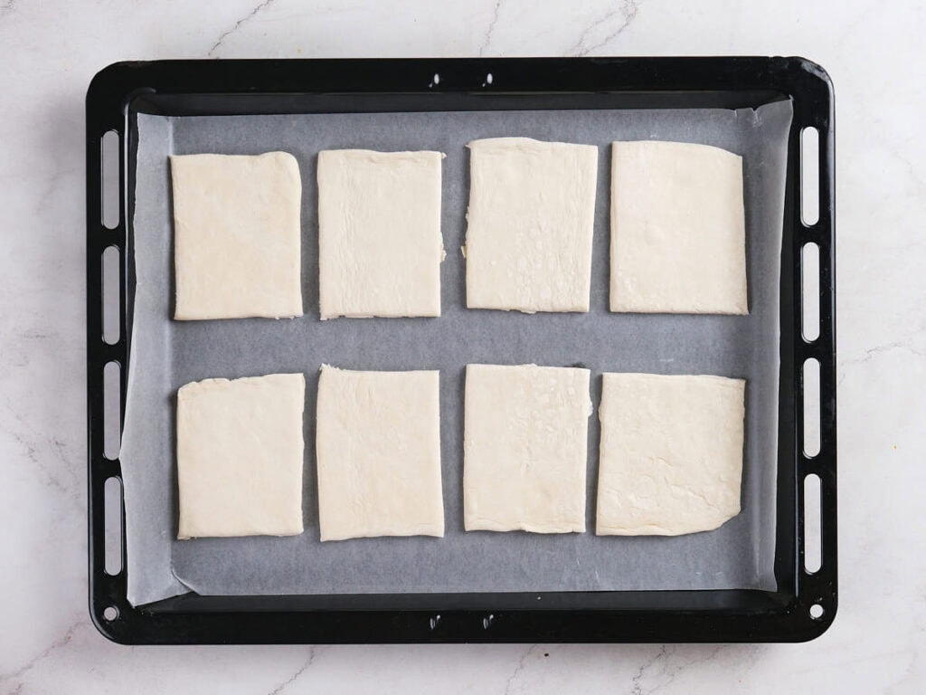 A black baking tray lined with parchment paper holds eight rectangular pieces of raw dough arranged in two rows.