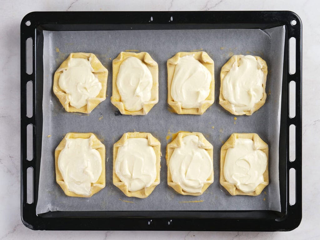 A baking tray with eight unbaked pastries topped with a creamy white filling, arranged on parchment paper.