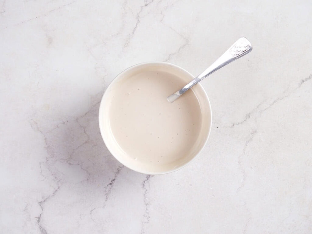 A bowl of creamy white sauce with a spoon resting inside, placed on a light marble surface.