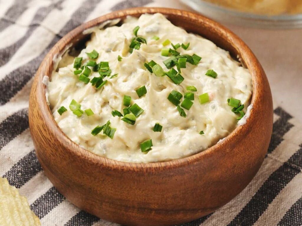 A wooden bowl filled with creamy dip topped with chopped green onions, resting on a striped cloth.