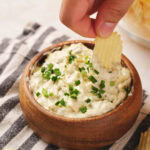 A hand dips a crinkle-cut chip into a wooden bowl of creamy chip dip topped with chopped green herbs, set on a striped cloth.
