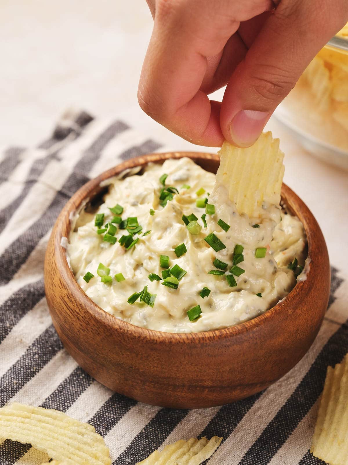 A hand dips a crinkle-cut chip into a wooden bowl of creamy chip dip topped with chopped green herbs, set on a striped cloth.