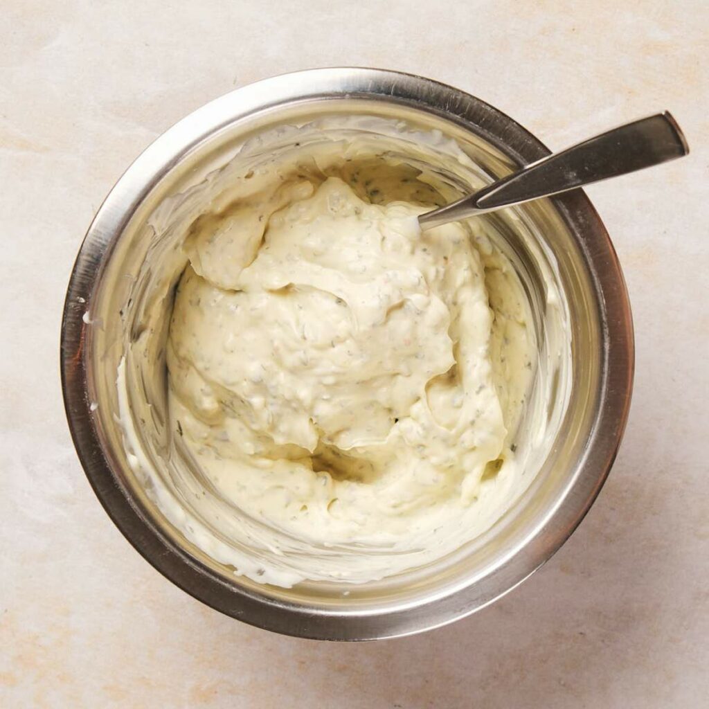 A metal bowl containing creamy white dip with herbs, and a spoon inside, placed on a light surface.