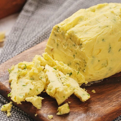 A loaf of garlic butter on a wooden board, partially sliced, with a gray cloth underneath.