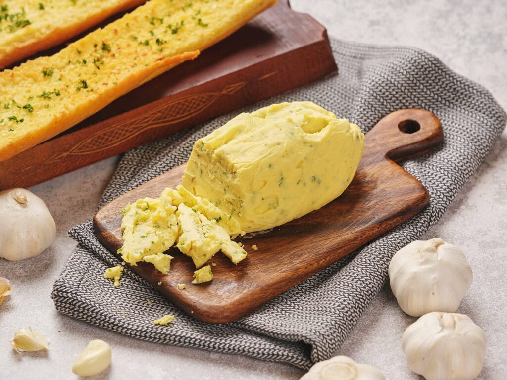 A block of garlic butter sits on a wooden board next to whole garlic bulbs. Behind it, garlic bread is displayed on a wooden tray.