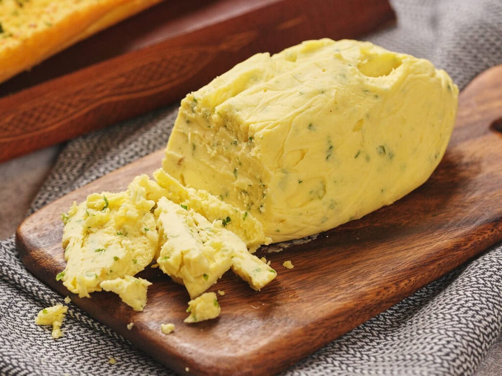 A block of garlic butter with several slices cut off, resting on a wooden cutting board with a gray cloth underneath.