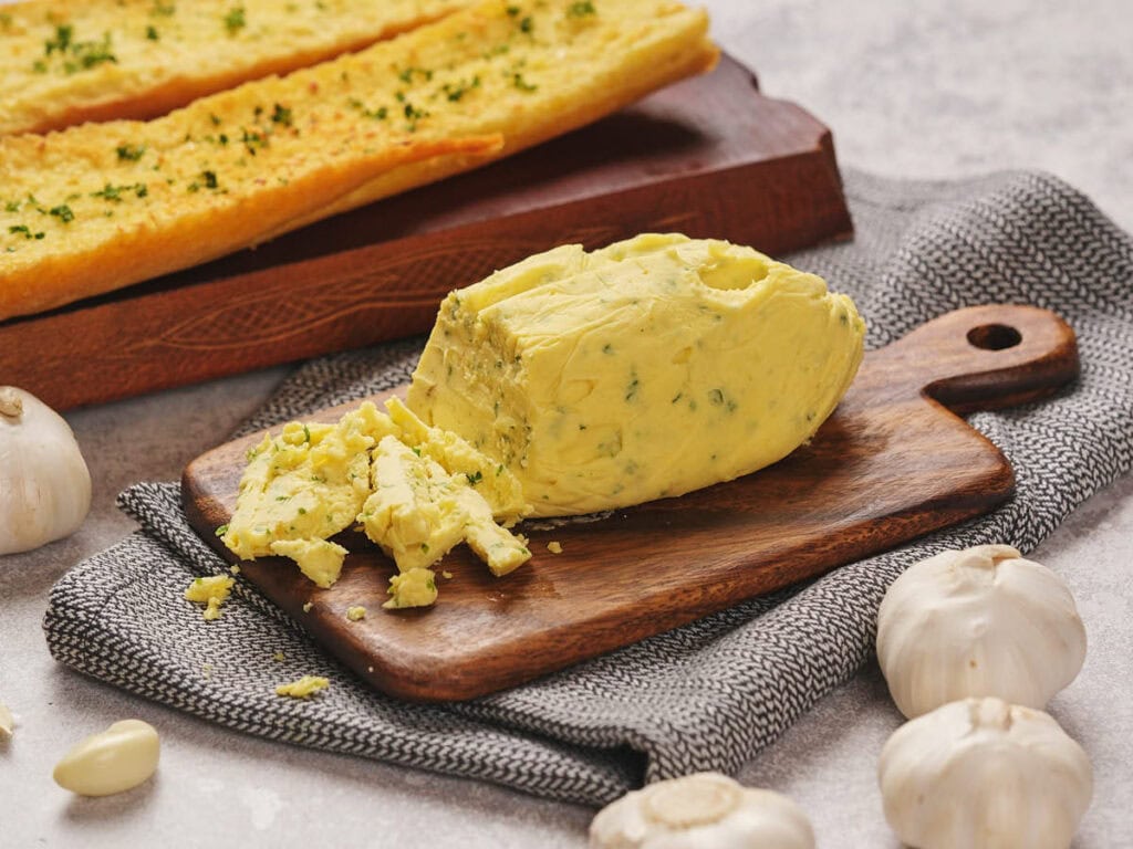 A block of garlic butter partially sliced on a wooden board with a gray cloth.