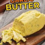 A block of garlic butter with herbs sits on a wooden board, partially sliced, next to a textured gray cloth.