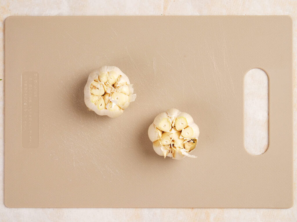 Two halved garlic bulbs placed on a rectangular, beige cutting board.