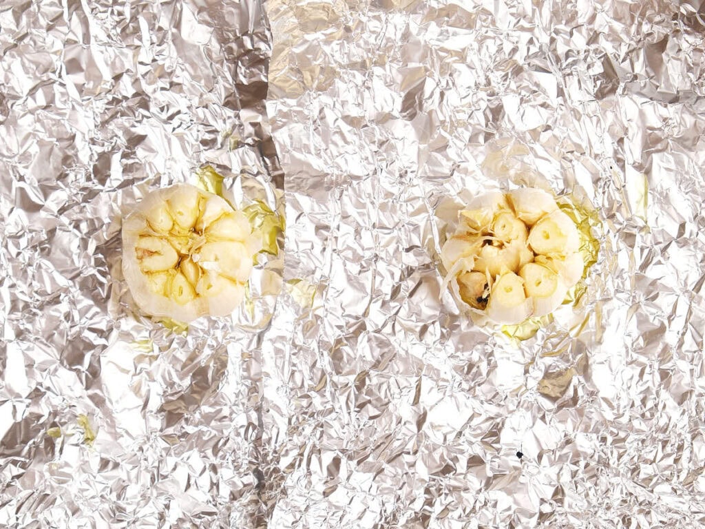 Two garlic bulbs cut in half and placed on crumpled aluminum foil.