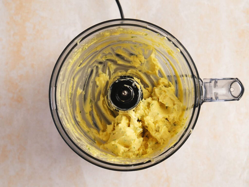 Overhead view of a food processor bowl containing a mixture of yellow butter and herbs, with some residue on the sides, set on a light countertop.