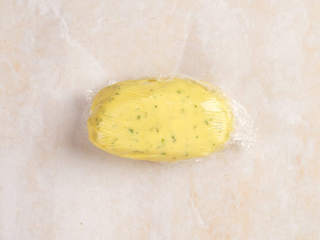 A ball of yellow dough with green herbs is wrapped in clear plastic wrap, placed on a light marbled surface.